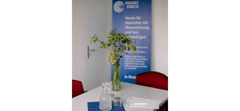 Ein Tisch. Darauf liegen: leere Blätter mit einem Stift, eine Glaskaraffe und zwei Gläser und eine Pflanze in einer Vase. Im Hintergrund steht ein Werbe-Rollup von FRAGILE Zürich.