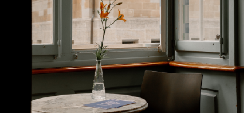 Eine Blumenvase auf einem Tisch. Dort liegen ebenfalls blaue Flyer. Im Hintergrund ist ein geöffnetes Fenster.