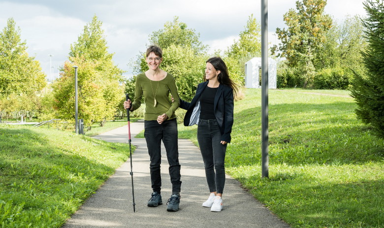 Zwei junge Frauen laufen auf einer Strasse. Eine davon hat eine Gehbehinderung.