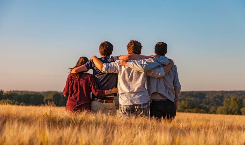 Vier Personen stehen in einem Weizenfeld mit dem Rücken zur Kamera. Sie legen ihre Arme um die Schultern der Personen neben ihnen.