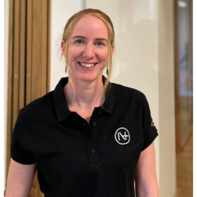 Profilfoto einer Frau mit hellen Haaren. Sie trägt ein schwarzes Poloshirt mit einem Logo darauf und lächelt in die Kamera.