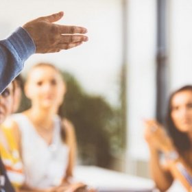 Unscharfes Bild von mehreren Personen an einem Tisch, die eine Person im Vordergrund anschauen. Von dieser ist nur der Arm zu sehen, sie macht eine Geste mit der Hand.