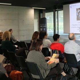 Une femme se tient derrière un pupitre, des personnes assises face à elle l'écoutent