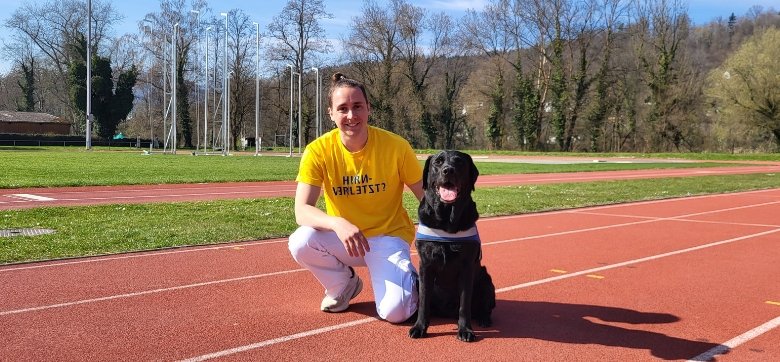 Ein junger Mann mit gelbem T-Shirt kniet auf einer Laufbahn, ein schwarzer Labrador sitzt neben ihm.