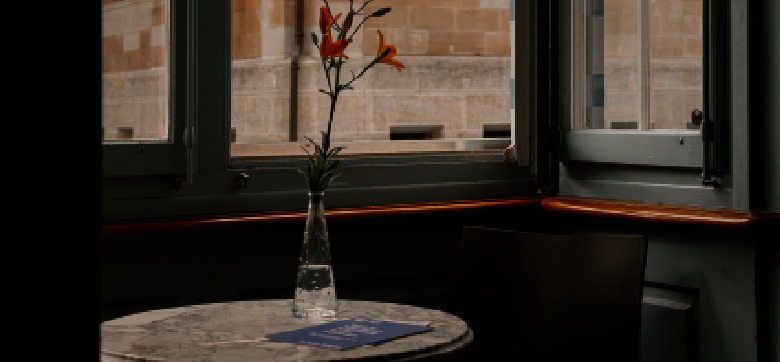 Eine Blumenvase auf einem Tisch. Dort liegen ebenfalls blaue Flyer. Im Hintergrund ist ein geöffnetes Fenster.