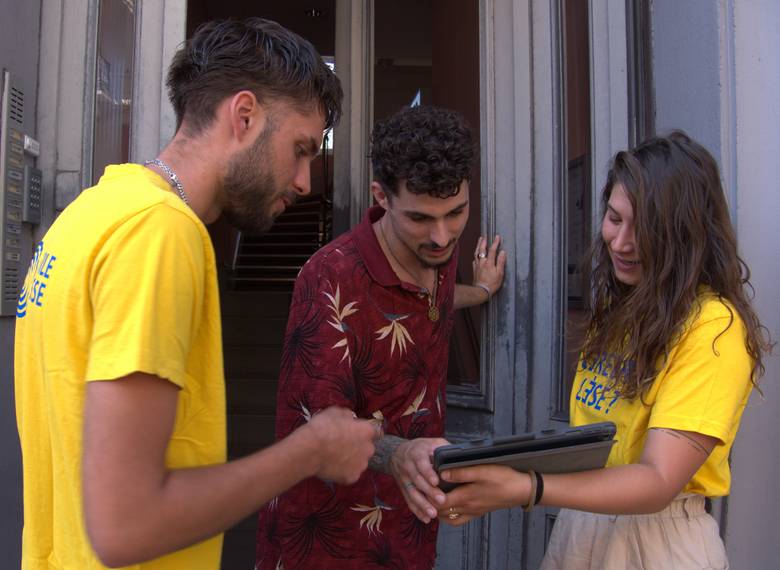 Zwei junge Menschen mit gelben T-Shirts sprechen mit einem Mann an der Haustüre.
