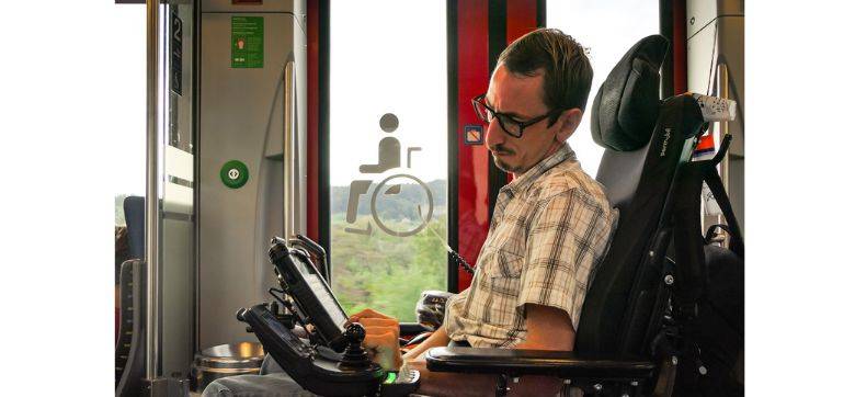 Ein Mann im Rollstuhl ist im Zug und bedient ein elektronisches Hilfsmittel.