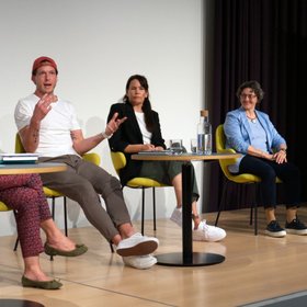 Fünf Personen sitzen verteilt an drei Tischen auf einer Bühne.
