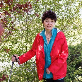 Eine Frau mittleren Alters mit kurzen, dunklen Haaren blickt in die Kamera. Sie trägt eine blaue Bluse und eine rote Jacke. In der rechten Hand hält sie einen Stock. Sie ist von grünen Sträuchern und Pflanzen umgeben.