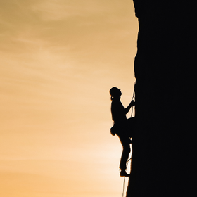 Silhouette einer Person, die eine steile Felswand heraufklettert, vor einem Sonnenaufgang.