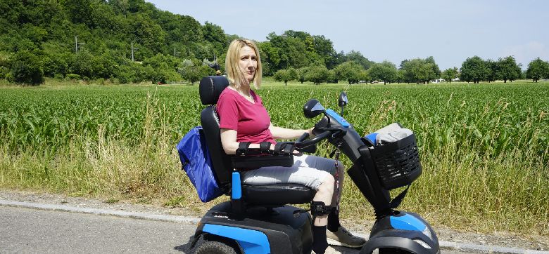 Eine Frau im elektrischen Rollstuhl, draussen auf einer Strasse vor einem Feld
