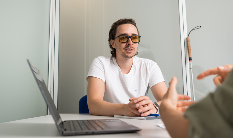 Ein junger Mann mit Sonnenbrille hört einer Person ihm gegenüber zu. Neben ihm stehen ein PC und ein Blindenstock.