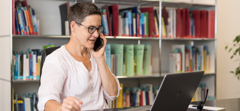 eine Frau sitzt vor ihrem Laptop und hält ein Smartphone an ihr Ohr.